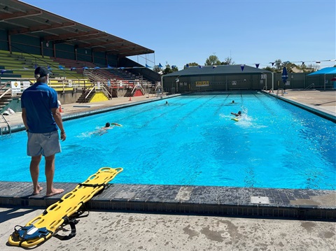 Lifeguards carrying out safety drills swimming at cessnock swimming pool. A man standing at the edge of a pool with a stretcher at his feet.